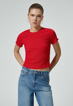 Rotes, kurzes T-Shirt mit strukturiertem Design, kurzen, fransigen Ärmeln, kombiniert mit hoch geschnittenen blauen Jeans. Deutliche Passform und Farbkontrast.