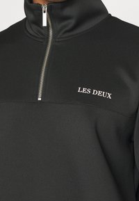 Sudadera de forro polar negra con media cremallera y textura suave, que presenta una cremallera plateada y un logo blanco de "LES DEUX" en el lado izquierdo del pecho.