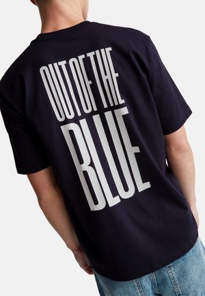Personne portant un t-shirt noir avec un grand texte blanc "OUT OF THE BLUE" dans le dos, associé à un jean bleu.