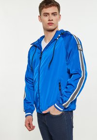 Giacca a vento in nylon blu con cappuccio, con strisce bianche e grigie sulle maniche, chiusura frontale con zip e tasche laterali.