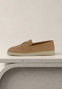 Pantof slip-on plat, culoare tan, cu talpă albă și o curea cu un mic detaliu metalic, expus pe o suprafață de marmură deschisă la culoare.