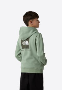 Mintgrønn hettegenser med en stor hvit logo og tekst på baksiden, med kengurulomme og ribbestrikkede mansjetter. Brukes med svarte bukser.