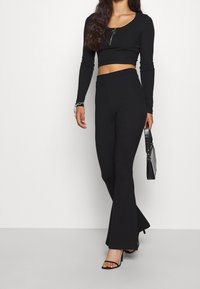 Schwarzes, verkürztes Langarm-Oberteil mit Reißverschlussdetail, kombiniert mit schwarzen, ausgestellt geschnittenen Hosen und Riemchensandalen. Eine kleine, strukturierte schwarze Handtasche rundet den Look ab.