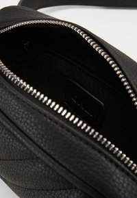 Musta teksturoitu nahkainen olkalaukku auki, näyttää sisäisen vetoketjutaskun ja metalliset vetoketjuhampaat aukon reunalla.