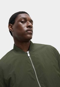 Giacca bomber verde oliva realizzata in materiale leggero, con zip frontale e colletto a coste, dotata di una finitura liscia e leggermente textured.