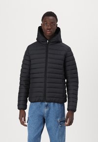 Doudoune noire avec capuche, présentant des panneaux matelassés horizontaux, une fermeture éclair à l'avant et des poignets élastiques, accompagnée de jeans bleus.