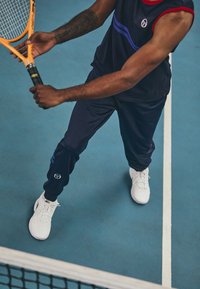 Mornarsko športno oblačilo z rdečimi in modrimi detajli, ki vključuje brezrokavnik in tekaške hlače, v kombinaciji z belimi teniškimi čevlji in oranžno loparjem.