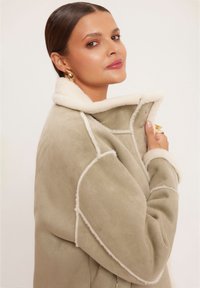 Zielona zamszowa kurtka z białym kołnierzem i mankietami z owczej skóry, z detalami w postaci guzików i kontrastowym przeszyciem wzdłuż szwów.