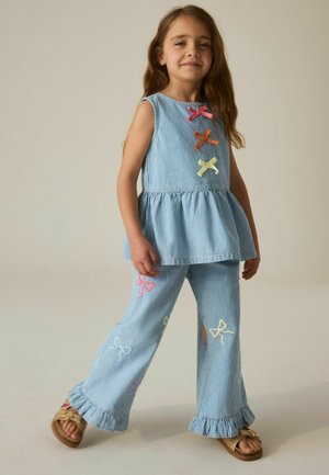 Young girl wearing blue and white striped sleeveless top and pants with colorful bow appliques and gold sandals, standing with a slight smile.
