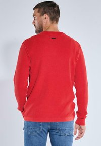 Red ribbed long-sleeve sweater with round neckline, featuring a small label on the back collar and fitted sleeves, paired with blue jeans.
