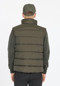 Olivenfarbene Pufferweste mit gesteppten Einsätzen, hohem Kragen und glatter, glänzender Textur. Getragen über einem dunklen Pullover, mit einer Mütze.