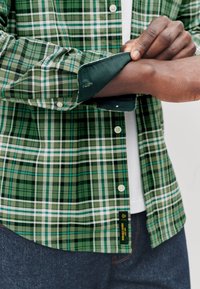 Green plaid shirt with varying shades, featuring buttons and a contrasting inner cuff. Cotton material with a smooth texture.