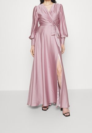 Vrouw die een lichtpaarse satijnen wikkeljurk draagt met lange pofmouwen, een ceintuur in de taille, een hoge split tot op de dij en zwarte open-toe hakken.