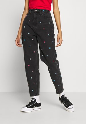 Person wearing black high-waisted jeans with scattered colorful letters, red top, white socks, and black low-top sneakers standing.