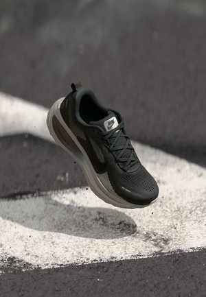 Chaussure de course noire avec semelle grise flottant au-dessus de l'asphalte avec des marquages routiers blancs, montrant le logo de la marque et des détails de conception en mesh.