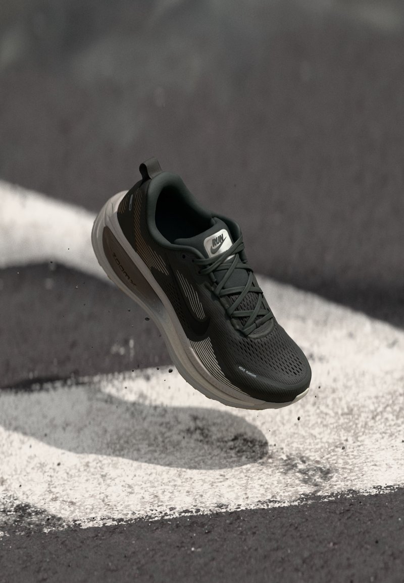 Chaussure de course noire avec semelle grise flottant au-dessus de l'asphalte avec des marquages routiers blancs, montrant le logo de la marque et des détails de conception en mesh.