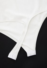Body in tessuto bianco con spalline regolabili, dotato di bottoni a pressione alla base e texture liscia, su uno sfondo nero.
