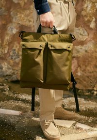 Nahrbtnik iz tkanine olivno zelene barve z dvema prednjima žepoma in črno opremo, opremljen z ročaji za nošenje in nastavljivimi trakovi. Teksturirana površina.