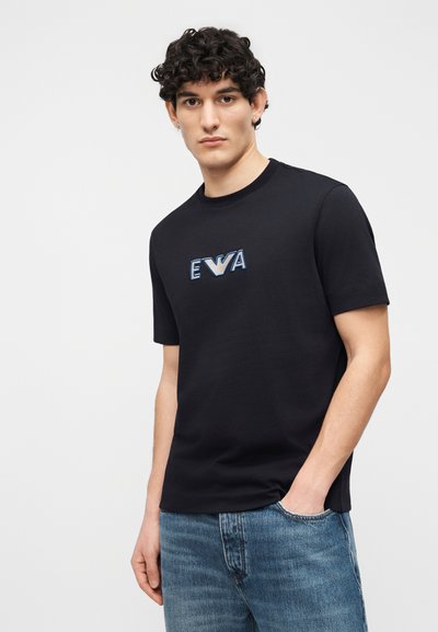 Νεαρός άνδρας με σγουρά σκούρα μαλλιά, που φοράει μαύρο μπλουζάκι με το λογότυπο "EWA" και μπλε τζιν, στέκεται με το ένα χέρι στην τσέπη μπροστά σε λευκό φόντο.
