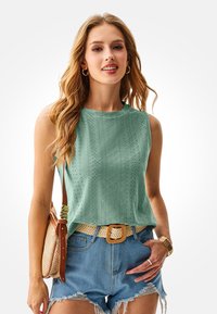 Light green sleeveless top with textured pattern, paired with denim shorts and a woven belt. The model carries a woven bag.