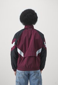 Corta-vento resistente à água em bordô e preto com detalhes em branco. Apresenta uma cintura elástica, colarinho alto e um ajuste solto.