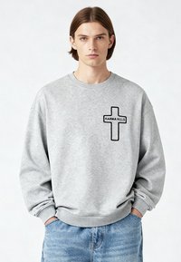 Jeune homme aux cheveux bruns raides portant un sweat-shirt gris avec une croix noire et le texte "KARMA KILLS", mains dans les poches d'un jean bleu.