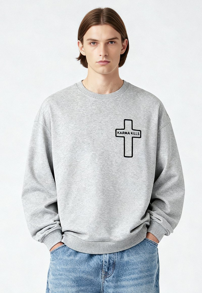Jeune homme aux cheveux bruns raides portant un sweat-shirt gris avec une croix noire et le texte "KARMA KILLS", mains dans les poches d'un jean bleu.