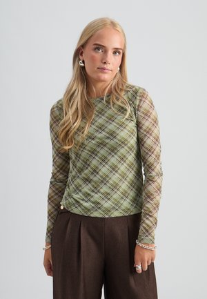 Femme blonde portant un haut à manches longues à carreaux verts et un pantalon taille haute marron foncé avec des bijoux en argent, debout devant un fond uni.