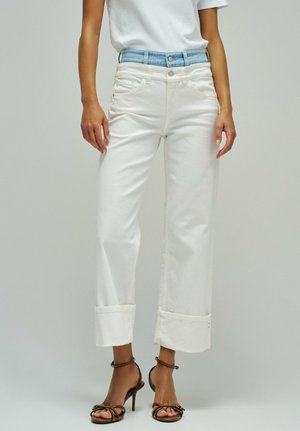 Femme portant un jean blanc à jambes larges avec de grands revers, un détail de ceinture en denim bleu clair, un t-shirt blanc et des sandales à talons hauts marron.