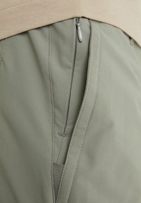Gros plan d'une fermeture éclair partiellement ouverte sur un pantalon vert olive porté avec un haut beige, montrant la texture du tissu et les détails des coutures.