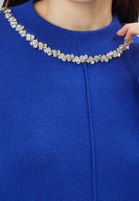 Maglione blu con un collo a coste decorato con un motivo di chiari strass luccicanti. Tessuto liscio e design aderente.