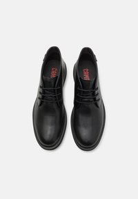 Zapatos de cuero negro con un acabado suave, puntera redonda y diseño de cordones. Presentan una plantilla acolchada con el nombre de la marca visible en rojo.