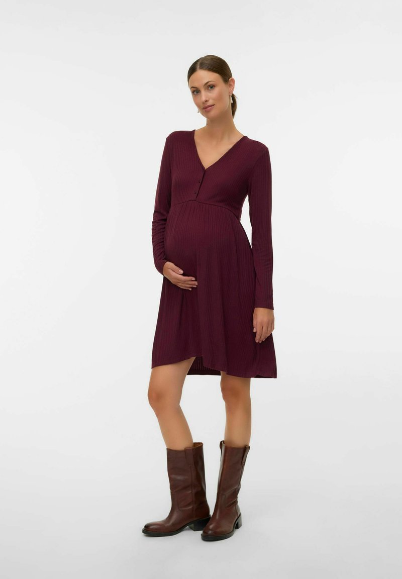 Robe de maternité en bordeaux foncé, texture côtelée, décolleté en V, manches longues, longueur genou, associée à des bottes en cuir marron hauteur genou.