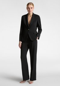 Completo su misura nero con un unico bottone dorato. Presenta un profondo scollo a V, pantaloni diritti e una texture del tessuto liscia.