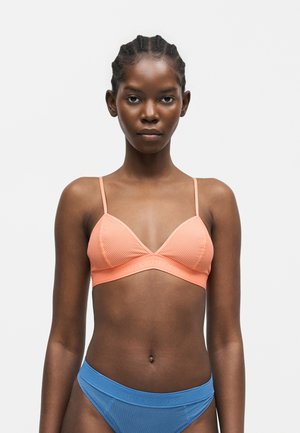 Žena nosi ribovaný broskyňový podprsenku s tenkými ramienkami a ribované modré bikiny spodky na jednoliatom bielom pozadí.