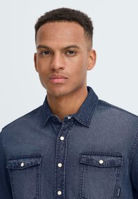 Denim shirt in donkerblauw met knoopsluiting aan de voorkant, twee borstzakken en witte knopen. Gelaagde stof met zichtbare naden.
