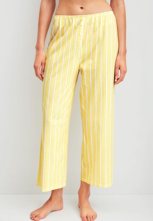 Frau trägt gelbe Schlafanzughose mit weißen und hellblauen vertikalen Streifen, steht barfuß vor hellem, einfarbigem Hintergrund.