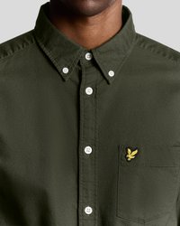 Camisa de botones verde oliva con cuello puntiagudo, botones blancos y un pequeño logo amarillo bordado en el bolsillo izquierdo del pecho.