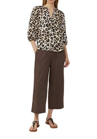 Bluse mit Leopardenmuster, Knopfverschluss vorne und 3/4-Ärmeln, kombiniert mit locker sitzenden braunen Hosen und schwarzen Flip-Flops.