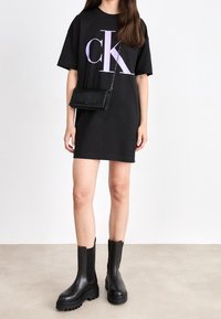 Czarna bawełniana sukienka t-shirtowa z dużym fioletowym logo 'CK', w połączeniu z małą fakturowaną czarną torebką i butami na grubej podeszwie w kolorze czarnym.