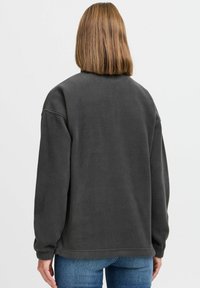 Felpa in pile grigia scura con spalle scese e polsini a coste, caratterizzata da una texture morbida e una vestibilità rilassata. La vista posteriore mostra un design semplice.