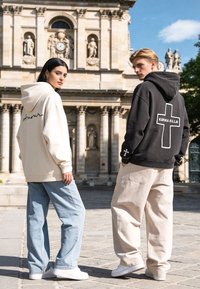 Deux jeunes adultes se tiennent à l'extérieur devant un bâtiment historique, portant des sweat-shirts à capuche avec des motifs d’écritures et de croix, associés à des pantalons larges et des baskets.