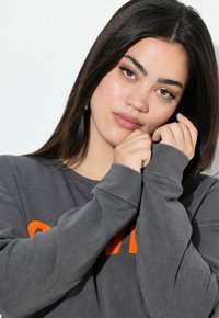 Sudadera gris de manga larga con letras naranjas, con un cuello redondo y un corte holgado. La tela parece suave y ligeramente texturizada.