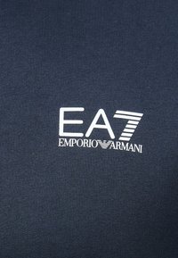 Námornícka modrá textília s bielym logom; "EA7" a "EMPORIO ARMANI" vytlačené tučným písmom s horizontálnymi čiarkami. Hladká textúra.