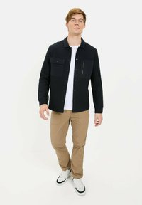 Navy quilted shirt jacket with two chest pockets, a zip pocket, and a button-up design, paired with beige pants and white sneakers.