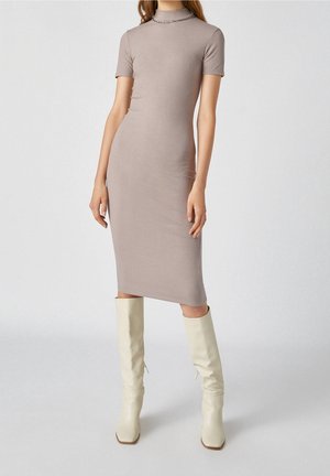 Femme portant une robe midi taupe ajustée en maille côtelée avec manches courtes et col montant, associée à des bottes écrues jusqu'aux genoux.