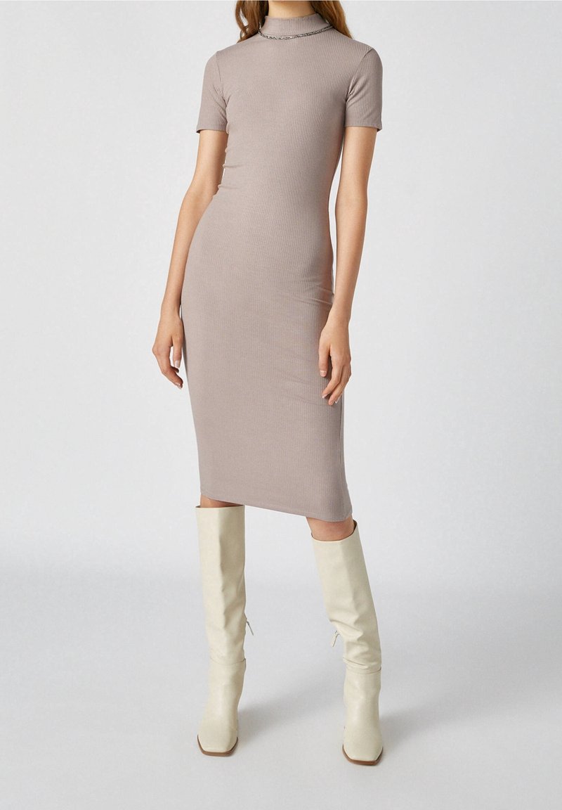 Femme portant une robe midi taupe ajustée en maille côtelée avec manches courtes et col montant, associée à des bottes écrues jusqu'aux genoux.