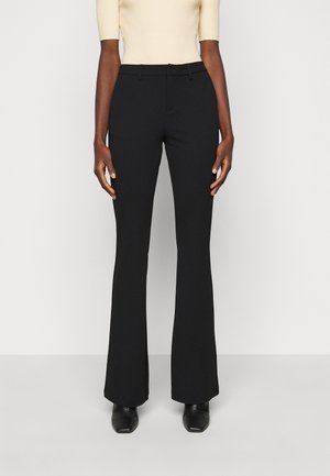 Personne portant un pantalon noir évasé taille haute, un haut beige clair à manches courtes rentré, et des chaussures noires à bout carré, debout devant un fond uni.