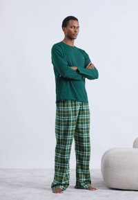 Πράσινο πουκάμισο με μακριά μανίκια συνδυασμένο με παντελόνια πιτζάμας σε πράσινο και κίτρινο καρό σχέδιο; απαλό ύφασμα και χαλαρή εφαρμογή. Στέκεται σε ένα ανοιχτό γκρι χαλί.