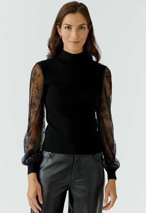 Pull noir à col montant, avec des manches longues en dentelle noire transparente. Le corps est en tricot, tandis que les manches présentent des détails en dentelle florale.